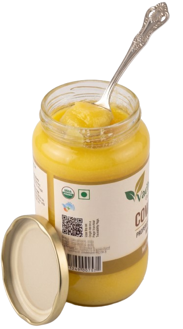 A glass jar of yellow ghee with its lid off and a decorative spoon resting inside, shown on a plain white background.